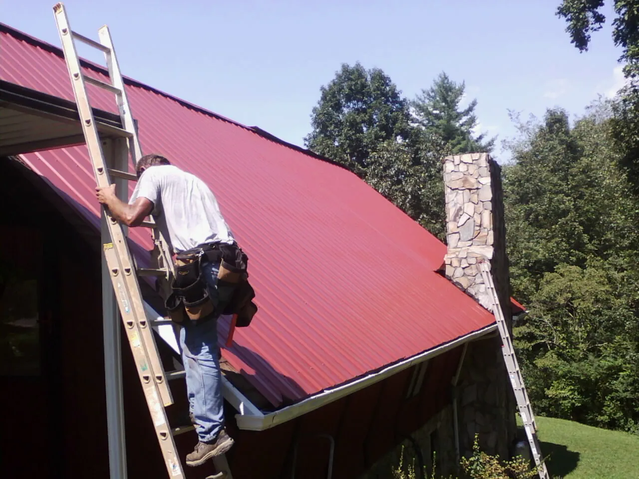 Roofer on a ladder beside a red metal roof and stone chimney — Soffit & Fascia Repair by Roofing Brothers in Tappan